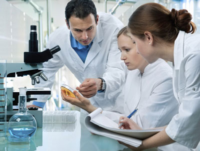 Three people in lab coats looking at a pitri dish next to a microscope